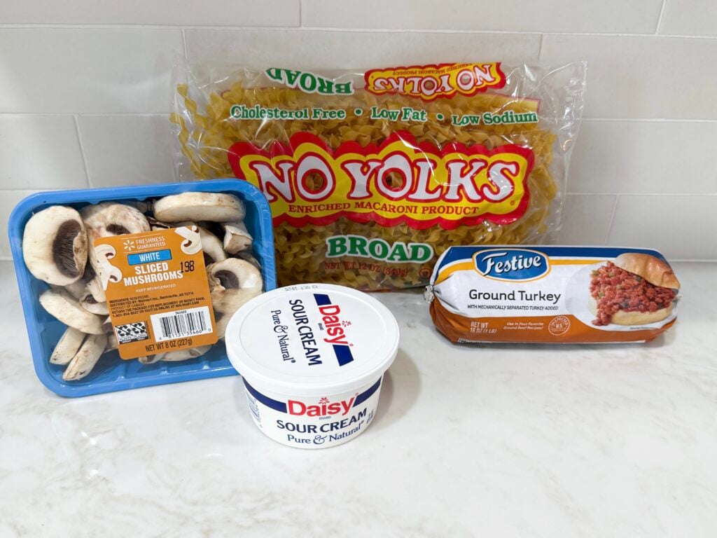 Turkey stroganoff ingredients with egg noodles, fresh mushrooms in a package, sour cream, and ground turkey all on a white counter