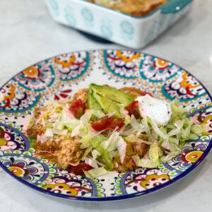 Pan of cheesy red chili chicken enchiladas topped with tomatoes, avocado, lettuce, and sour cream.