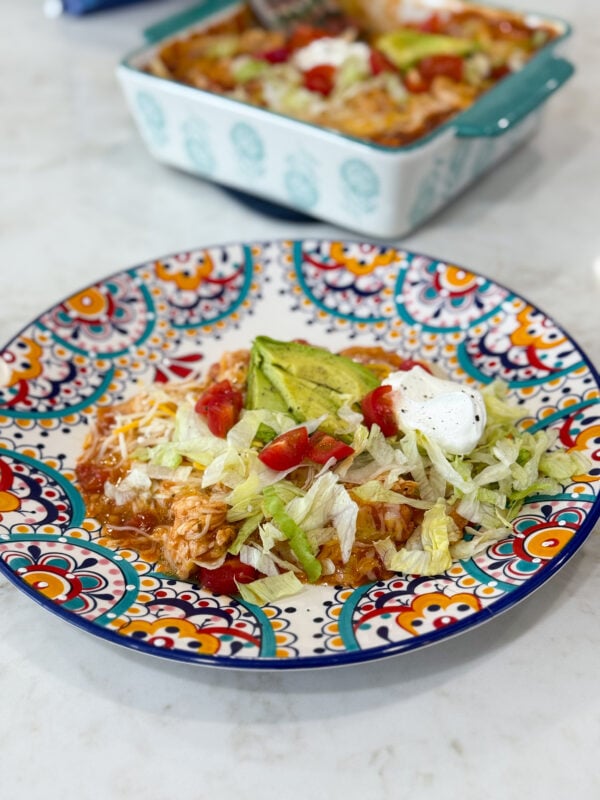 Pan of cheesy red chili chicken enchiladas topped with tomatoes, avocado, lettuce, and sour cream.