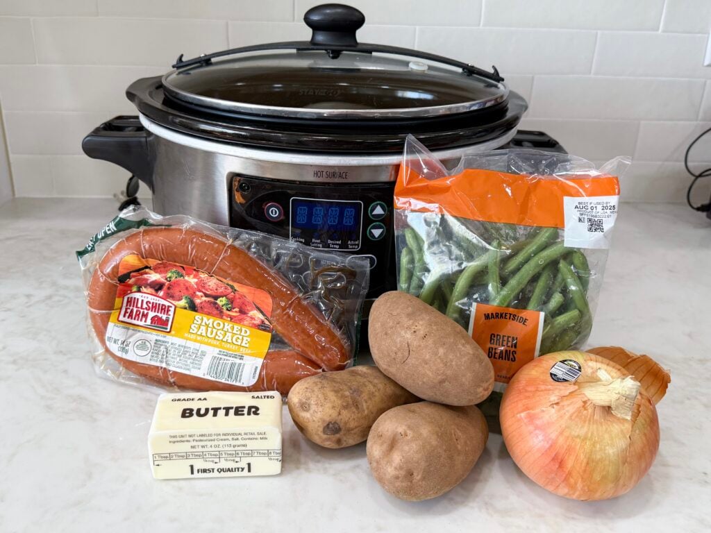 large black and grey slow cooker with sausage, green beans, onion, butter, and potatoes on a white counter