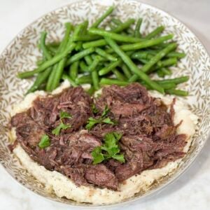 Shredded beef over mashed potatoes with green beens on a grey and white plate