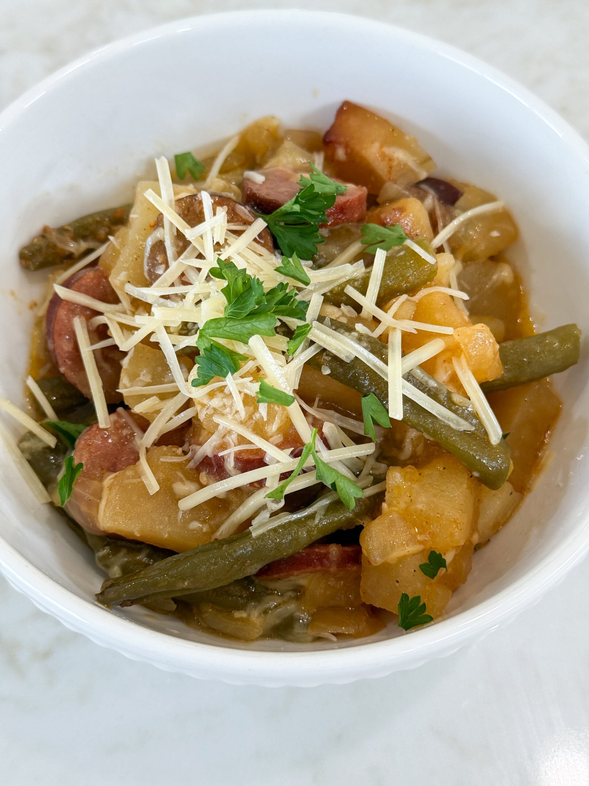 Cooked sausage, potatoes, and beans in a white bowl topped with parmesan cheese and fresh parsley