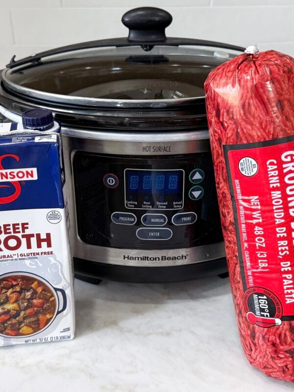 large slow cooker on a white counter with beef broth and package of ground beef